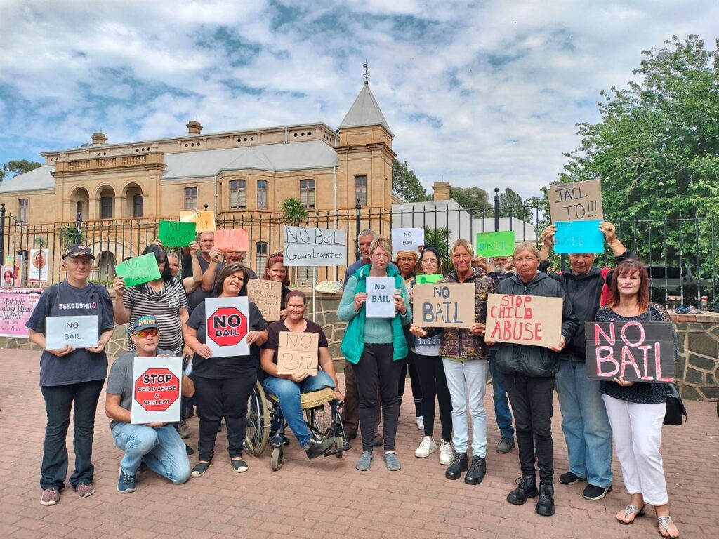 Groep mense voor hof by hofverskyning van vier mense wat in aanhouding in in verband met aanranding van kinders.