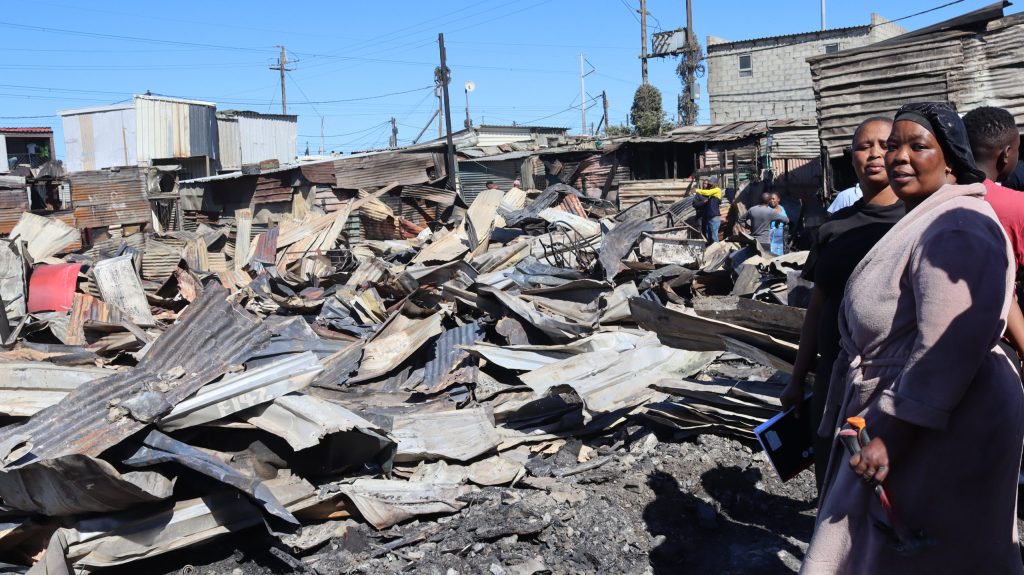 Residents stand among fire debris in Ethembeni settlement with blackened wood and metal sheets scattered around