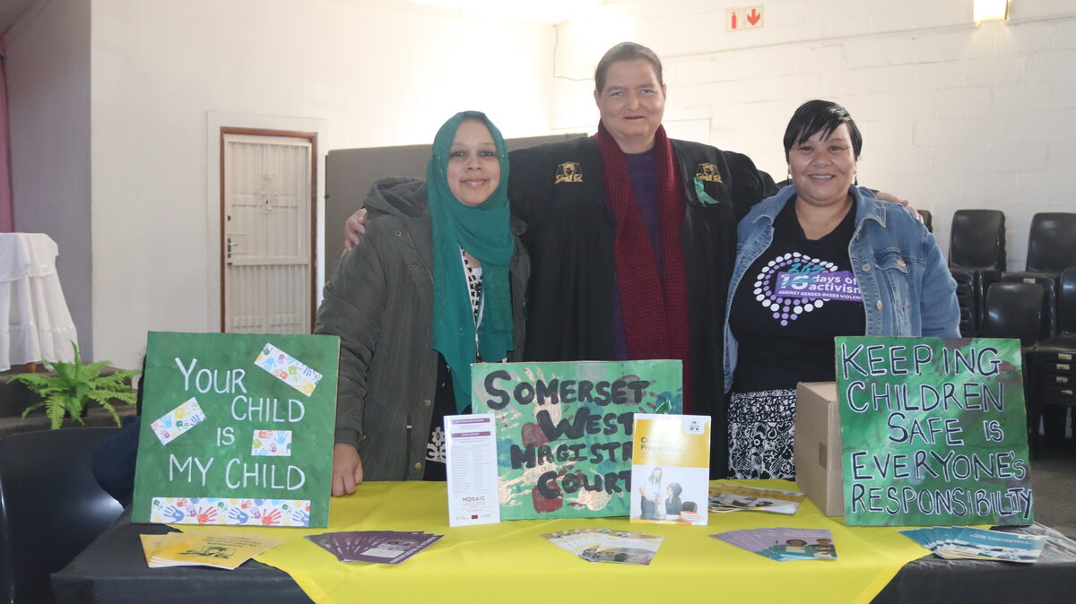 Tasneem Seboa, Ursula Sansen and Clarina Ward stand beside a child protection awareness board at the Strand event.