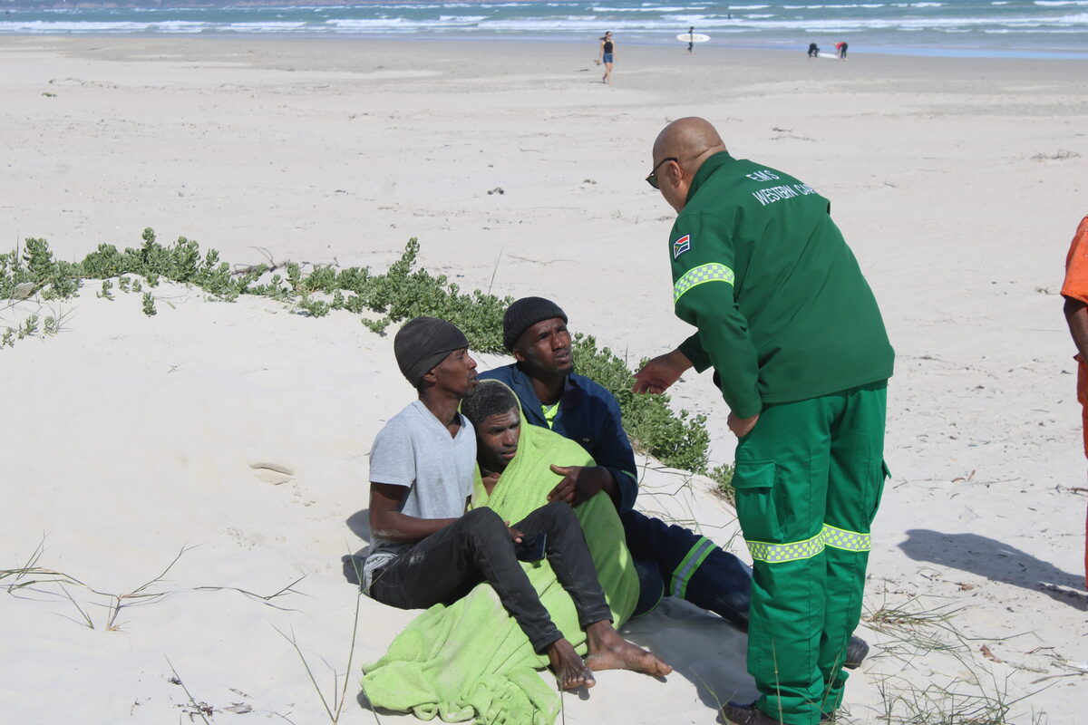 A Western Cape EMS paramedic attends to the rescued swimmer, wrapped in a blanket on the dunes, as companions look on. Search efforts continue for two other swimmers reported missing in the surf at Strand Beach. Photo: Yaseen Gaffar
