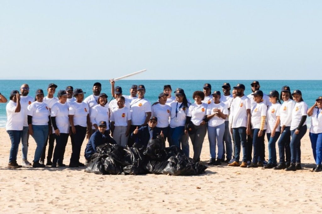 A large group of people standing with black bags at the beach.