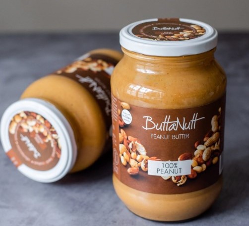 ButtaNutt peanut butter jar label showing the brand name "ButtaNutt" with "PEANUT BUTTER" and "100% PEANUT" text prominently displayed on the packaging.