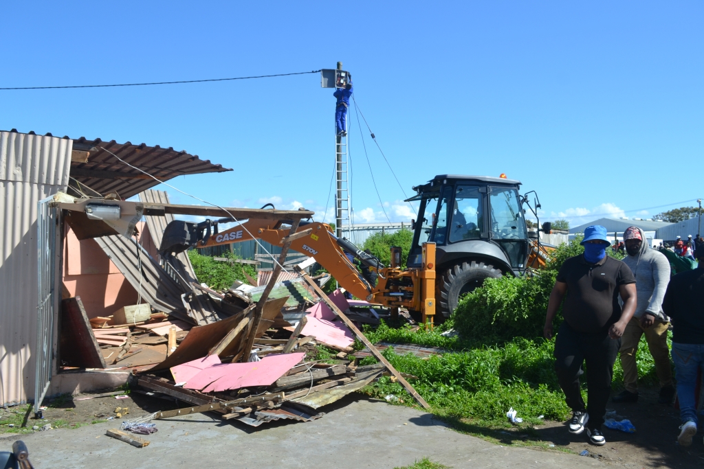 A bulldozer demolishes an informal structure in Airport Valley as residents look on.