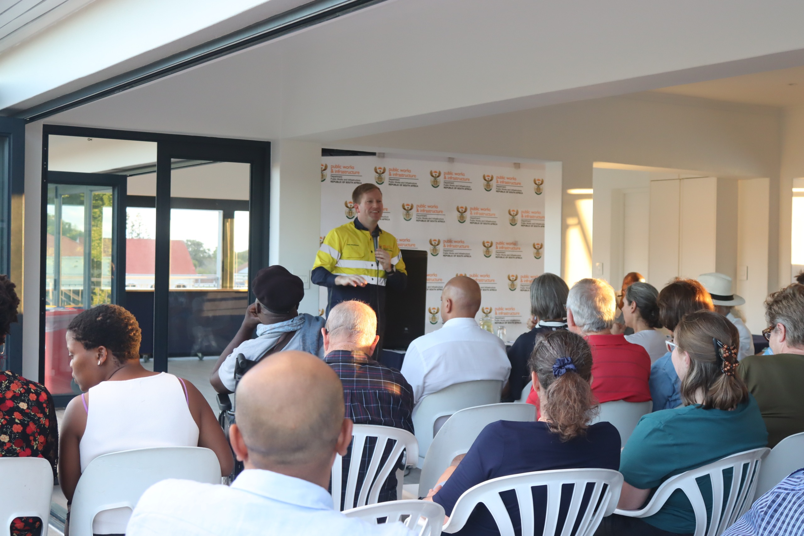 Public Works Minister Dean MacPherson stands at the front of a packed hall at Wynberg Boys’ Junior School, addressing seated residents during a community meeting about delays and security concerns at the Waterloo Green property.