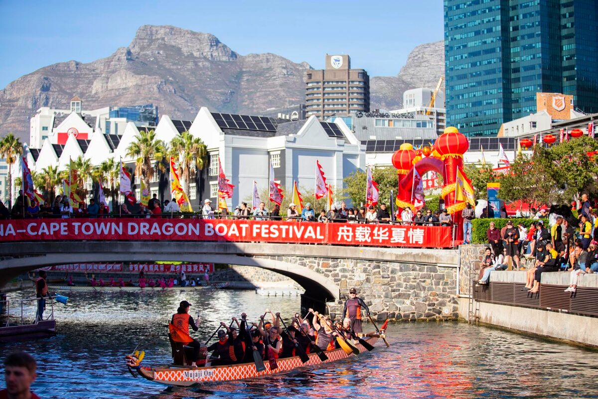 A dragon boat crew paddles beneath a festival banner as spectators line the banks of the waterway, with Table Mountain providing a dramatic backdrop to the racing action.