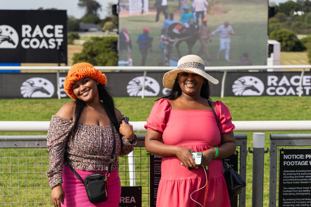 Two smiling women pose in front of a racecourse fence wearing a beanie and a straw hat.