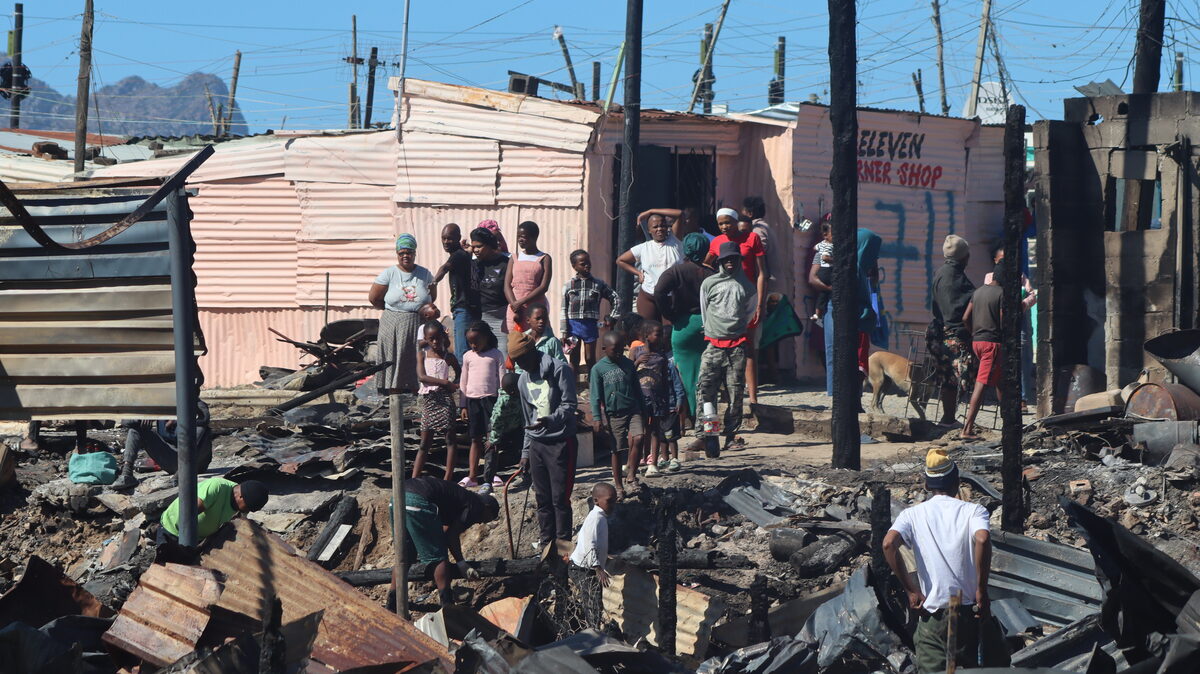 Residents overlooking charred remains in Ethembeni settlement with scattered debris and blackened timber