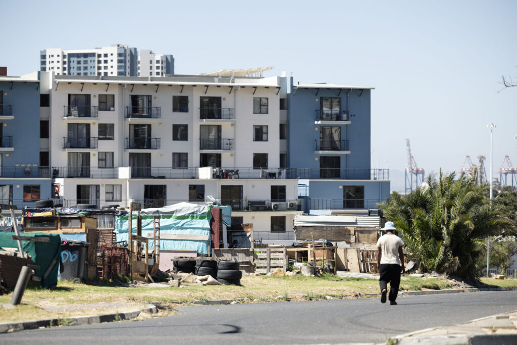 A man walk past a homeless informal settlement situated next to an apartment buidling in downtown Cape Town on December 18, 2025. Nestled between spectacular mountains and the Atlantic Ocean, Cape Town has emerged as a growing tourism destination, topping in 2025 the Telegraph and Time Out magazine's rankings of best cities in the world. Activists say the drive to profit from tourism accommodation is pricing locals out of coveted neighbourhoods, including areas near District Six -- a site long associated with the apartheid-era removals that displaced non-white communities to remote, underdeveloped townships. (Photo by GIANLUIGI GUERCIA / AFP)