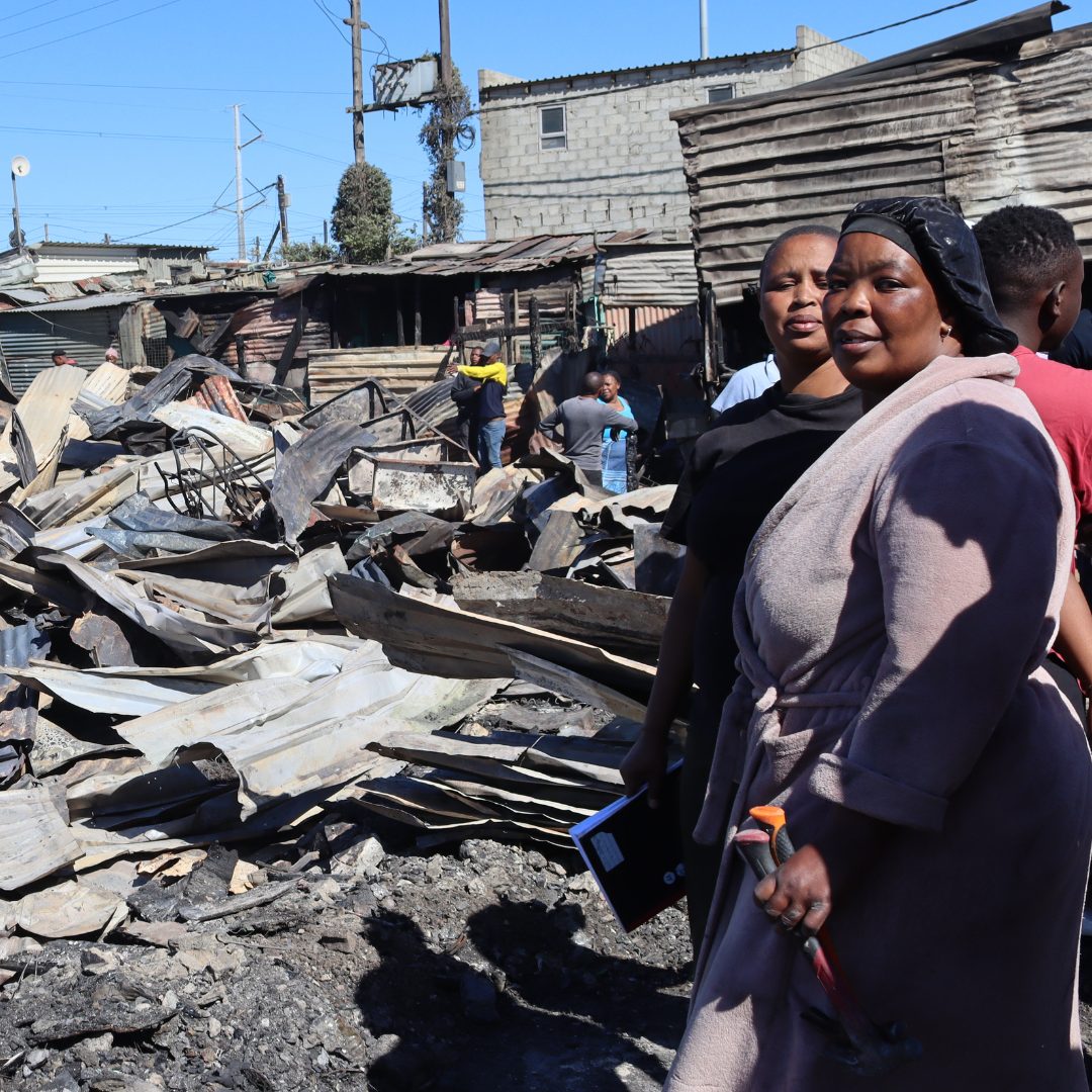 Residents stand among fire debris in Ethembeni settlement with blackened wood and metal sheets scattered around