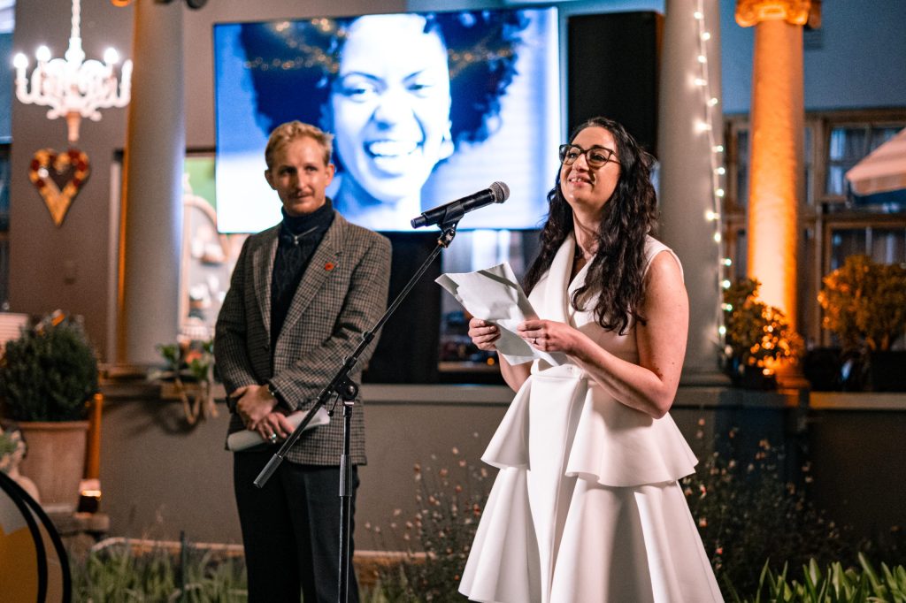 Michael Garbett (feesdirekteur) staan en toekyk hoe Marijda Kamper (programbestuurder) die Chrystal Donna Roberts-beurs op die programbekendstelling van die 2026-fees bekend stel. 'n Foto van Roberts is agter op die skerm sigbaar. FOTO: ZitaFilmz