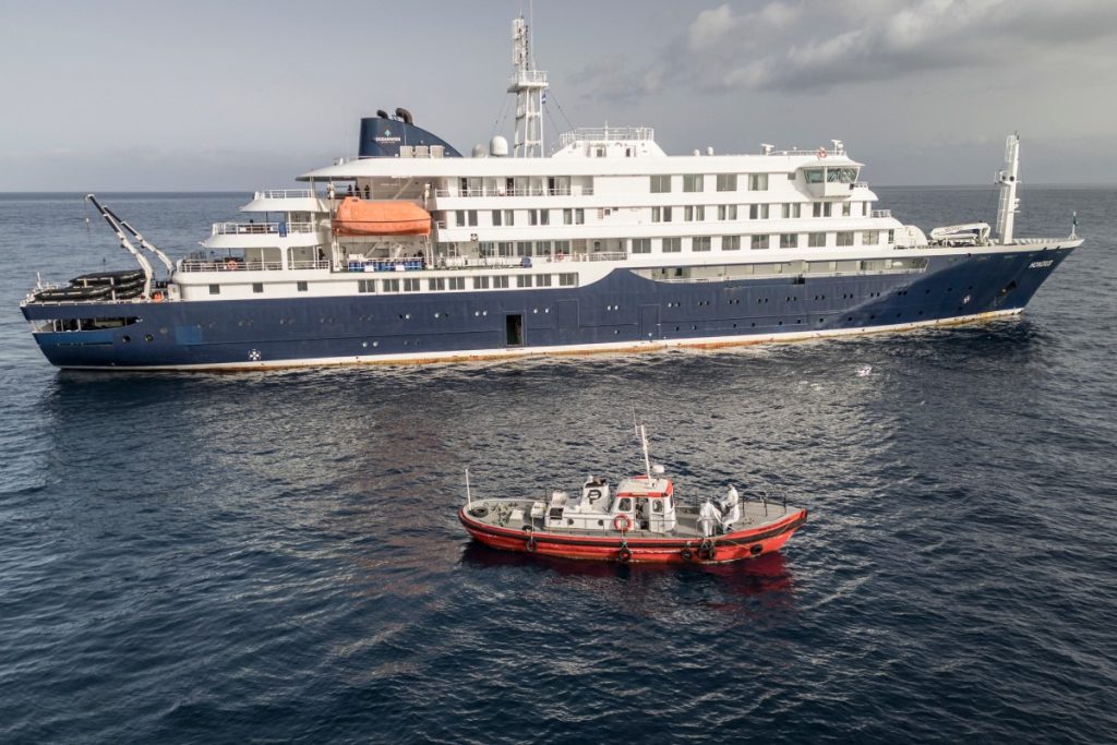 An aerial view of an ambulance boat carrying crew members wearing hazmat suits as they approach the pilot door on the starboard side of the cruise ship MV Hondius, while stationary off the port of Praia, the capital of Cape Verde, on May 5, 2026. Two seriously ill crew members on a cruise ship stricken by a deadly hantavirus outbreak will be evacuated via Cape Verde to the Netherlands, allowing the vessel to sail on to Spain's Canary Islands, the ship operator said Tuesday. The MV Hondius has been at the centre of an international health scare since Saturday, when WHO was informed that the rare disease -- usually spread from infected rodents typically through urine, droppings and saliva -- was suspected of being behind the deaths of three of its passengers. As others fell ill, passengers and crew have been in isolation after Cape Verde authorities barred the ship from docking, and as health authorities scrambled to find a port that would take the Hondius. (Photo by AFP)