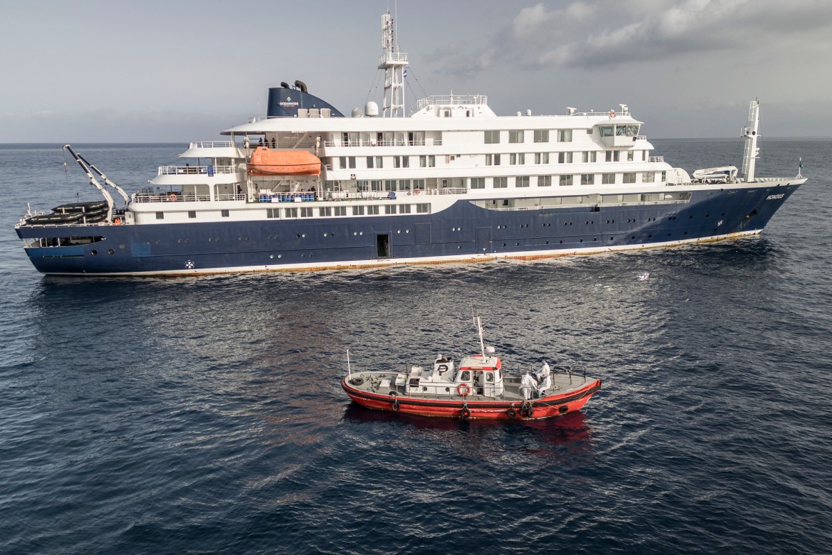 An aerial view of an ambulance boat carrying crew members wearing hazmat suits as they approach the pilot door on the starboard side of the cruise ship MV Hondius, while stationary off the port of Praia, the capital of Cape Verde, on May 5, 2026. Two seriously ill crew members on a cruise ship stricken by a deadly hantavirus outbreak will be evacuated via Cape Verde to the Netherlands, allowing the vessel to sail on to Spain's Canary Islands, the ship operator said Tuesday. The MV Hondius has been at the centre of an international health scare since Saturday, when WHO was informed that the rare disease -- usually spread from infected rodents typically through urine, droppings and saliva -- was suspected of being behind the deaths of three of its passengers. As others fell ill, passengers and crew have been in isolation after Cape Verde authorities barred the ship from docking, and as health authorities scrambled to find a port that would take the Hondius. (Photo by AFP)