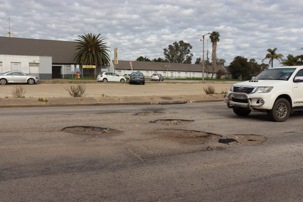 A pothole-riddled road outside the Uitenhage Train Station illustrates the deteriorating condition of roads in Kariega, with damaged sections spanning both lanes and posing a hazard to motorists.