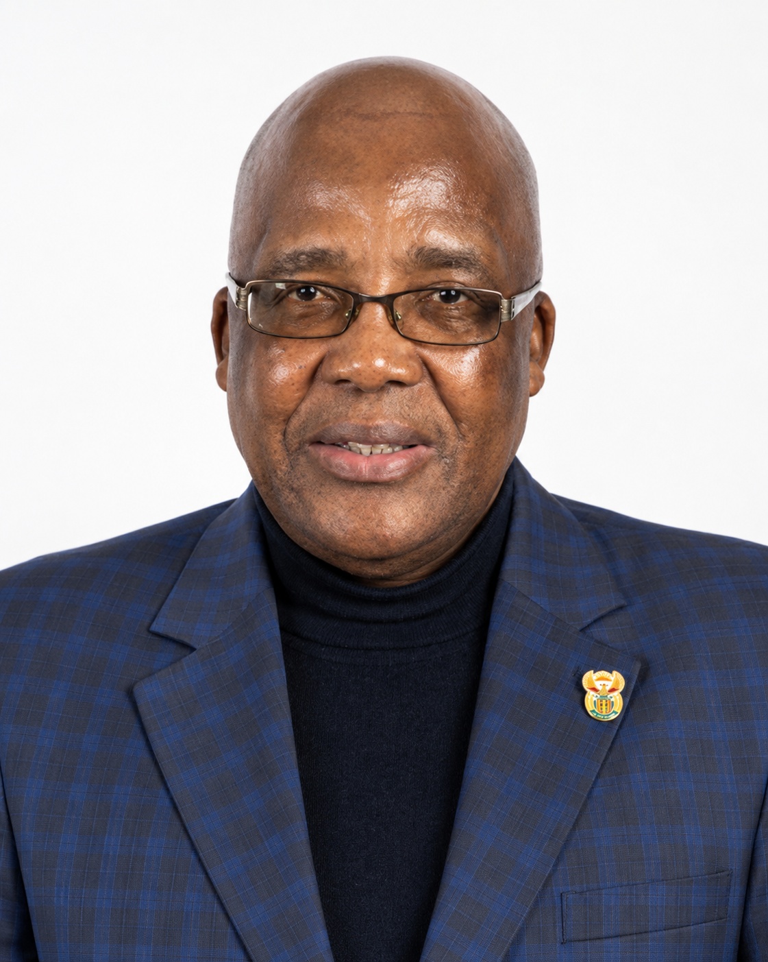 **Alt text:** > Health Minister Dr Aaron Motsoaledi, wearing a blue plaid blazer, navy turtleneck and rectangular glasses, poses for an official portrait against a white background. A small gold and green crest pin is visible on his lapel.
